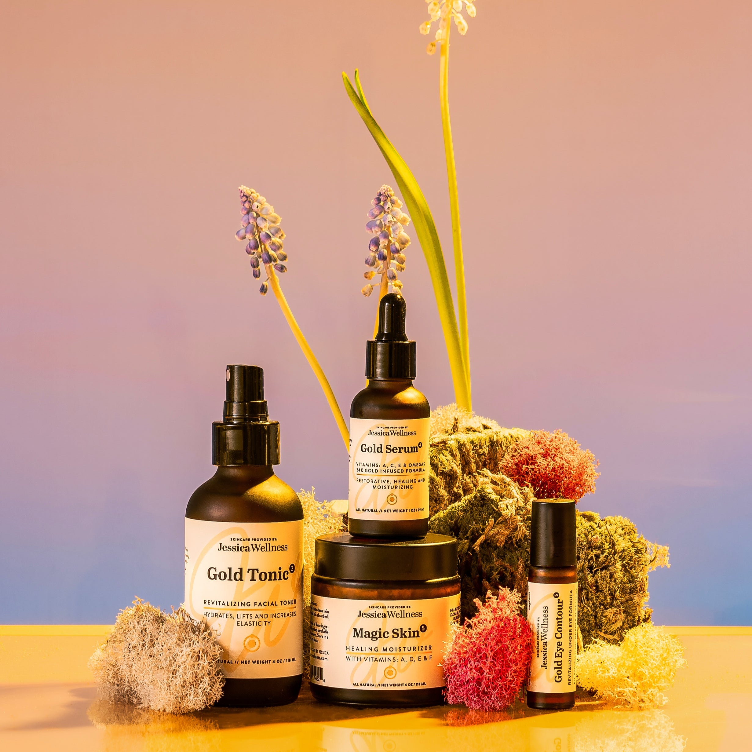 Four skincare products from Jessica Wellness, Gold Tonic, Magic Skin, Gold Serum, and Gold eye serum, are arranged on a reflective surface with soft moss and grape hyacinth flowers. The background features warm pink and golden tones, creating a calm, natural vibe.