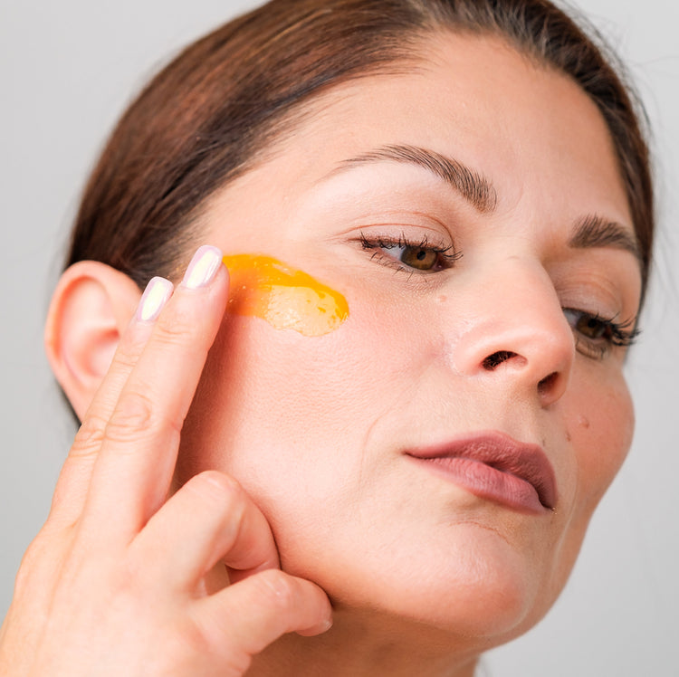 Woman applying a golden-coloured anti-aging serum to her cheek using her fingertips, showcasing skincare application and smooth, healthy skin.